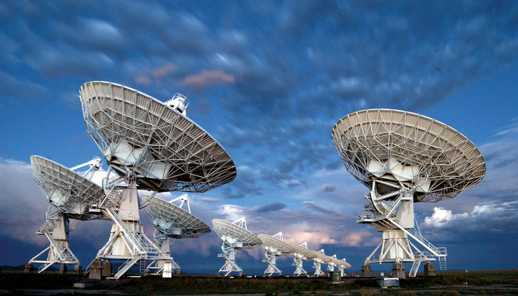 Dark wispy clouds over a row of massive radio telescope dishes stretching toward the horizon, their white paint standing out against the background in the fading light.