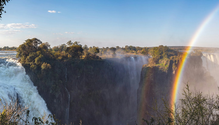 A set of three massive waterfalls surrounded by lush plant life and large trees basking in the sunshine. Water spray causes a vivid rainbow to appear against the cliffs and the pristine blue sky.